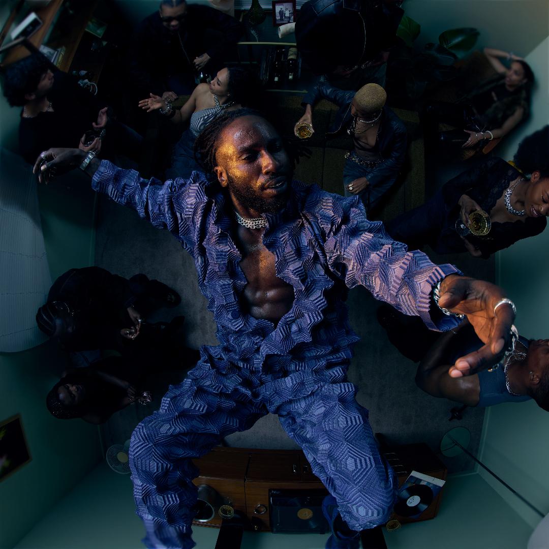 Man in a blue textured outfit surrounded by people in a room with a blue wall.