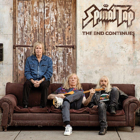 Three musicians sitting on a couch with a rustic door and 'Spinal Tap' album cover in the background.
